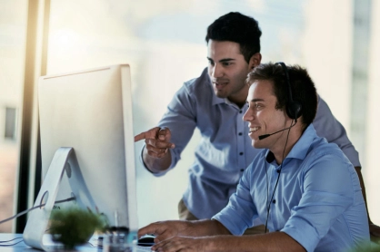 Two coworkers at a desk looking at a computer monitor; one seated wearing a headset, the other pointing at the screen.