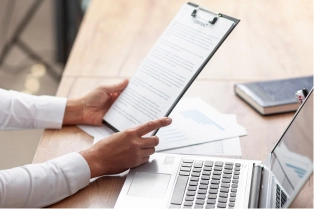 Person holding a clipboard with a document over a laptop and papers on a wooden desk in a workspace.